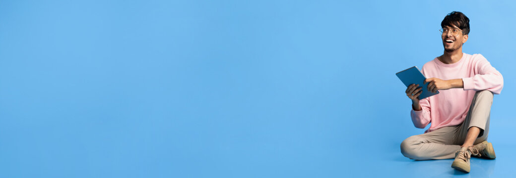 A young man with short dark hair and glasses sits cross-legged on the floor, smiling while holding a tablet. He wears a pink sweater and light pants in front of a solid blue background.