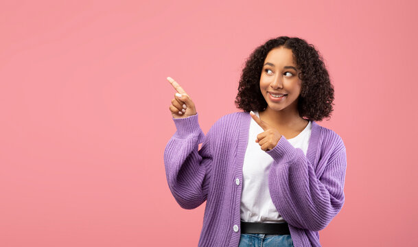 A cheerful young black woman stands against a soft pink backdrop, confidently pointing in two directions, inviting potential advertisements. She wears a relaxed outfit and a friendly expression.