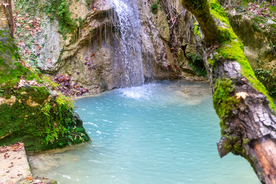 A small waterfall cascades down a rocky, mossy wall into a natural turquoise pool. Dense vegetation, vines, roots, and moss can be seen all around. (saklı, gizli cennet şelalesi) Manavgat - Antalya