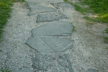 Old broken asphalt path with gravel pushed into the edges, flanked by green grass. Scene of ruins and abandonment, showing wear and neglect.