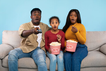 A young black family sits on a couch, shocked while watching a movie on TV. They are eating popcorn and the father points the remote at the screen. The blue background adds to the scene's liveliness.