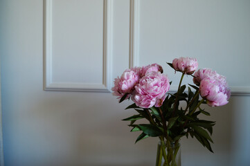 Pink Peonies in a Glass Vase Against White Wall. A bouquet of lush pink peonies with green leaves is arranged in a transparent glass vase set against a classic white wall with molding
