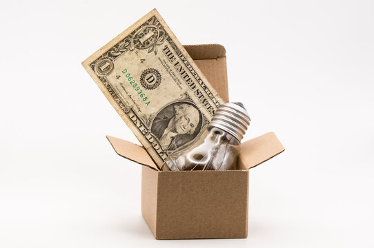 One dollar bill and an incandescent lightbulb standing upright in a small cardboard box, symbolizing the rising price of electricity, power, and utility bills.