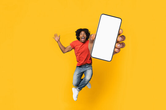 A young man in a red shirt is jumping with joy while holding a smartphone in front of a bright yellow background. His expression conveys excitement and energy.