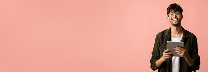 A young man dressed casually stands with a friendly smile, holding a tablet in his hands. The background is a soft pink color, creating a cheerful atmosphere.