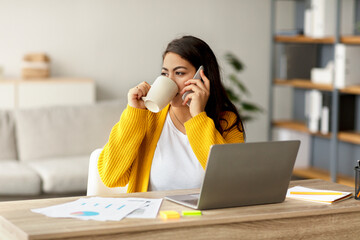 Arab woman in a living room talks on her smartphone while using a laptop. She sips coffee and works in a cozy home environment, illustrating a modern freelance lifestyle.