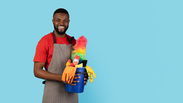 Cleaning Services. Handsome Black Cleaner Man Posing With Basket Of Detergents Over Blue Background, Smiling African American Male Janitor Ready For Spring Cleaning, Panorama With Copy Space - Powered by Adobe