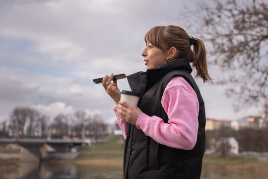 Young woman is sending a voice message on her phone while holding a takeaway coffee cup in a park, enjoying a moment of multitasking during a chilly day
