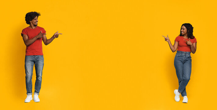 Two young adults in matching red shirts stand on a bright yellow background, playfully pointing at each other. They are smiling, creating a lively and energetic atmosphere together.
