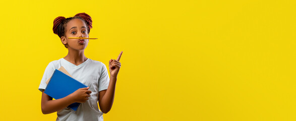 A child stands with a blue folder in one hand and a pencil in her mouth, looking thoughtful while pointing one finger up. The bright yellow backdrop adds a cheerful atmosphere.