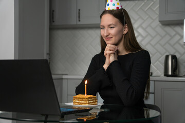 Young woman celebrating her birthday online, wearing a festive party hat while making a wish with a lit candle on a delicious cake, all set up in front of her laptop at home