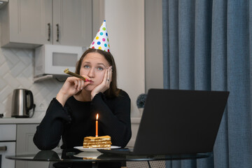 Young woman wearing party hat blowing in blowing a festive horn and looking bored while celebrating birthday online using laptop at home during lockdown