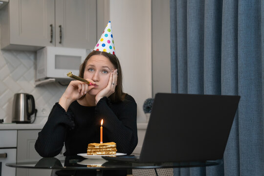 Young woman wearing party hat blowing in blowing a festive horn and looking bored while celebrating birthday online using laptop at home during lockdown