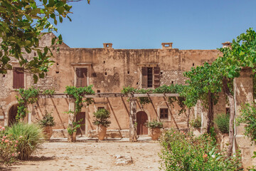 A rustic courtyard building of the Arkadi Monastery in Crete, Greece, featuring weathered stone walls, wooden doors, climbing plants, and warm Mediterranean colors. A notable cultural and historical l