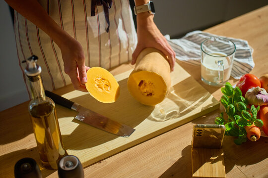 Person in apron holding sliced butternut squash on cutting board in kitchen. Woman slicing pumpkin on table with olive oil, spices, vegetables and smartphone. Concept of cooking and healthy food