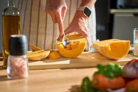 Close-up of woman removing seeds from halved pumpkin with spoon on wooden cutting board in kitchen. Person in apron preparing squash for roasting. Concept of home cooking and healthy food
