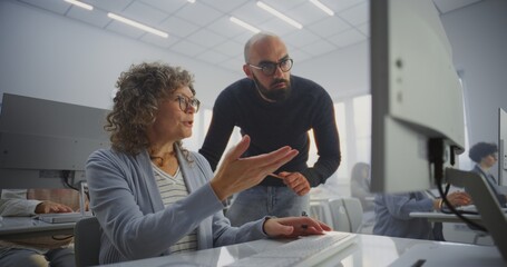 Middle Aged Female Student Works on Computer Project While Teacher Stands Beside, Guiding and Reviewing Progress. Concept Adult Education, Mentoring, Digital Learning, and Professional Development.