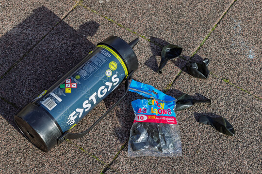 A disposable bottle of nitrous oxide (laughing gas) and several deflated latex balloons, abandoned on a textured pavement, depicting a discarded party element. Utrecht Netherlands, 10 November 2025.