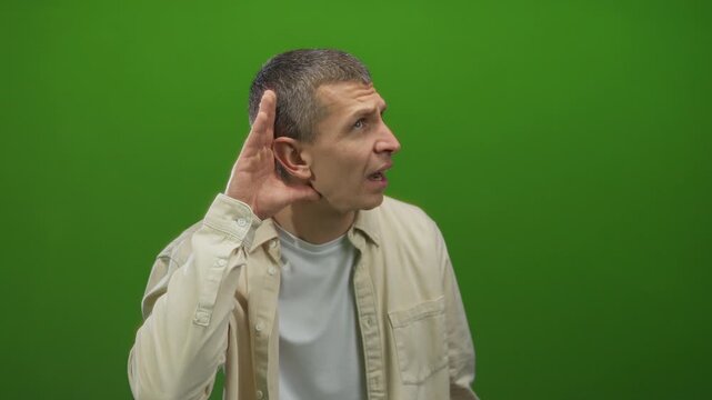 Man listening while holding hand to ear against vibrant green wall, conveying curiosity and concentration in an isolated environment, wearing casual white shirt emphasizing engagement.