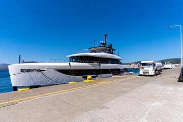 Luxury motor yacht being refueled the Marmaris marina pier on a clear sunny day, with a fuel tanker truck parked alongside the dock. Modern yacht design, waterfront marina view and harbor background.