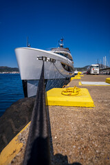 Vertical view of a luxury yacht moored at the marina pier with a strong mooring rope in the foreground and a fuel tanker truck in the background on a sunny day, showing modern yacht design and harbor 