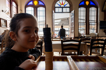 Young girl sitting in a traditional café playing with stacked backgammon pieces, with sunlight shining through stained-glass windows and people in the background, capturing a warm everyday scene.