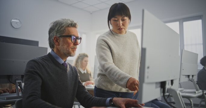 Teacher Assists Senior Student With Computer Project in Modern Classroom. Young Female Instructor Comments on Mans Project, Highlighting Details on Monitor. Digital Skills, Modern Adult Education.