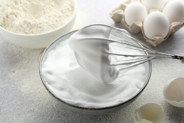 Beating egg whites in bowl, whisk, eggshell and flour on grey textured table, closeup