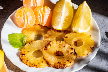 Grilled pineapple slices arranged with fresh mandarin segments and lemon pieces, creating a vibrant and refreshing tropical composition. The caramelized edges of the pineapple add warmth and texture, 