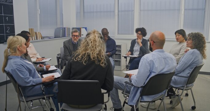 In Classroom With Large Windows and Chalkboard Schedule on Wall, Circle Adult Learners Listens Closely Facilitator. Everyone Seated on Chairs, Participating in Group Exercise or Guided Conversation.