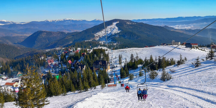 Active winter scene with people riding a ski lift over a snowy mountain slope overlooking a colorful alpine village and forested peaks under clear blue sky.