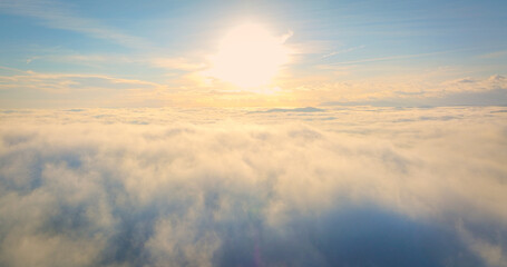 AERIAL: View above a layer of morning winter fog illuminated by bright sunlight. Soft mist hides the valley and only distant mountain peaks peek through fluffy clouds. Dreamlike, tranquil atmosphere.