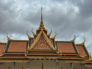 Fototapeta premium The ornate, gilded gable of a traditional Khmer temple roof at Royal Palace in Phnom Penh, Cambodia, stands in perfect symmetry against a vast, overcast, and cloudy sky, highlighting its fine detail.