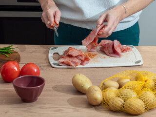 Woman cutting raw meat on a chopping board while preparing a homemade meal. Vegetables, potatoes and kitchen tools around. Natural home cooking setup.