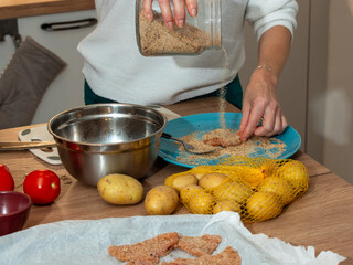 Close-up of a woman breading meat with breadcrumbs while preparing a homemade meal. Fresh ingredients like potatoes and tomatoes on the table. Authentic home cooking scene