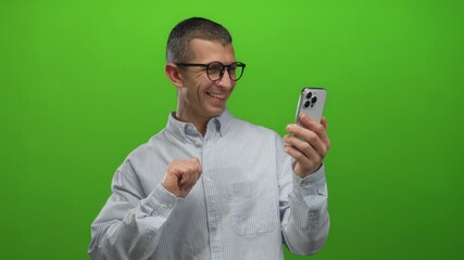 Man smiling at smartphone with green background engaging in video call showing cheerful expression and gesturing with hand while wearing glasses and casual shirt in isolated setting.