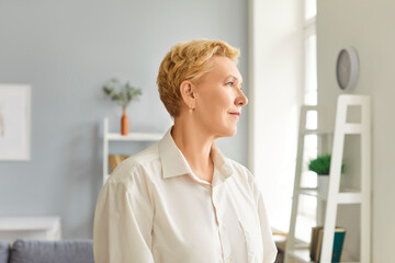 Portrait of mature blonde woman wearing light beige clothes looking dreamily at window with slight smile. Pensive senior lady looking into distance, lost in thought with peaceful expression at home.