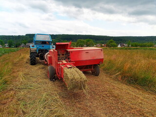 A blue tractor pulls a red hay baler machine, processing cut hay into bales in a rural field