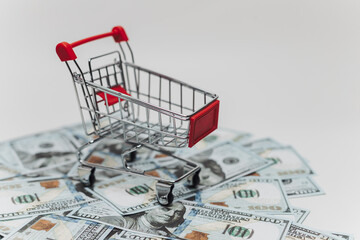 A miniature shopping cart rests atop a pile of US hundred-dollar bills against white background