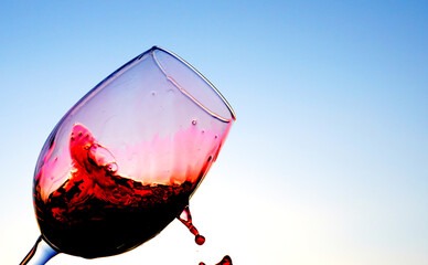 Red wine splash from glass on blue sky. Sommelier swirls with glass of red wine at sunset sky. Red Wine Swirling In Glass on vine Tasting, celebration party. Vibrant liquid splashing. Red splashes.