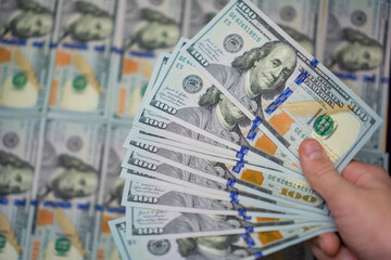 hand holding a fan of new one hundred dollar bills against a blurred background pattern of usa currency, concept of financial investment, banking transaction, business success