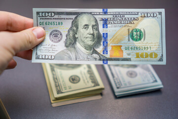 hand holding up a single one hundred dollar bill close up, over two neat stacks of cash on a dark surface, concept of financial audit, money inspection, cash transaction