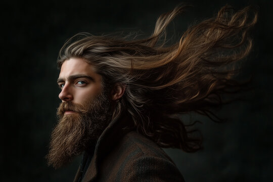 A man with long, flowing hair and a full beard looks over his shoulder against a dark background, with hair blowing in the wind.