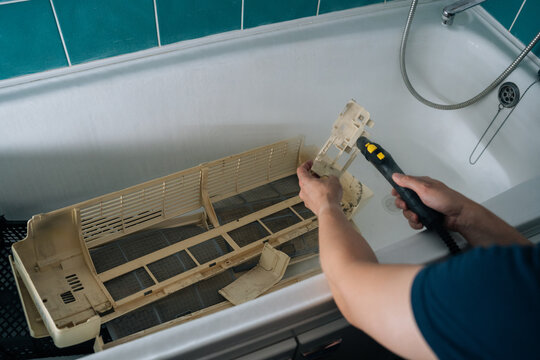 Close-up cropped shot of unrecognizable technician cleaning filter of air conditioner unit inside bathtub, utilizing steam cleaner to remove dirt and grime, ensuring optimal performance and hygiene.