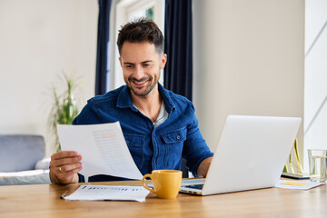 Smiling man looking at financial document working remote on laptop