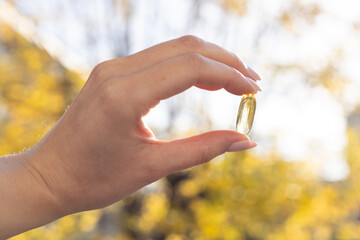 Closeup of a person holding a vitamin D 3 capsule against colourful trees in fall.