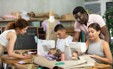 Experienced tailor supervising group of colleagues working together on custom order in sewing studio, monitoring garment assembly, checking fabric preparation, and reviewing drafted patterns..