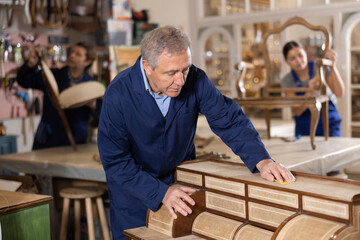 In restoration showroom, elderly craftsman carefully polishes tabletop of old chest of drawers with rounded drawers and twisted bronze handles with soft cloth