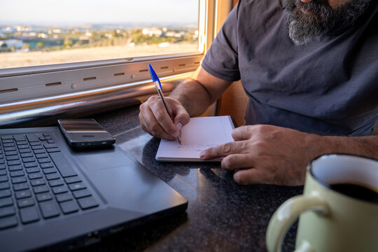 A middle-aged man writes in a notebook inside a camper van, with a laptop, phone, and coffee mug on the table, and a rural landscape visible through the window.