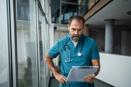 Male doctor in scrubs reading patient chart in hospital corridor